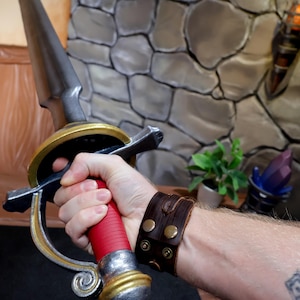 Puede incluir: Una espada con una hoja de color plateado, un mango rojo y una empuñadura de color dorado y plateado. La espada presenta una guarda curva y un pomo en la base del mango. El diseño incluye un acabado metálico en la hoja y la guarda.