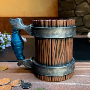 May include: A wooden-style beer mug with a silver handle and accents. The handle is wrapped in blue material. The mug has a brown, textured exterior with silver bands. A decorative item for a bar or home.