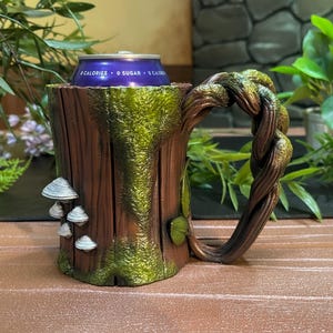 May include: A whimsical, handcrafted mug designed to resemble a tree stump. The brown and green mug features a twisted branch handle, faux mushrooms, and moss-like detailing. A purple can is inside the mug.