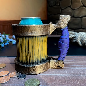 May include: A decorative beer stein with a gold-colored rim and handle, and a yellow and black body. The handle is wrapped in purple material. A blue can sits inside the stein. Coins are scattered on the wooden surface.