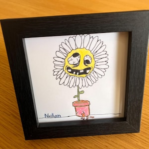 May include: A framed artwork featuring a cartoon flower with a yellow face and white petals. The flower is in a pink pot with a crack. The artwork is signed "Nathan" and is set in a black frame.