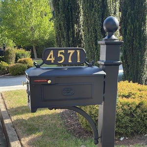 May include: Black mailbox with gold-colored numbers reading "4571" on a decorative sign. The mailbox has a classic design with a curved support arm and a gold-colored handle. The mailbox is mounted on a black post.