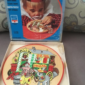 May include: A round wooden puzzle by Simplex Toys featuring a circus theme with a clown, a donkey, and a wagon labeled Circus Pipo. The puzzle includes small knobs on the pieces and comes in a blue box with a picture of a child playing with it.