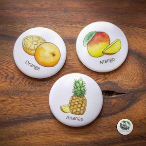 Op de afbeelding: Drie witte ronde knoppen met illustraties van fruit. De eerste knop toont een hele sinaasappel en een halve sinaasappel met het woord "Orange" eronder. De tweede knop toont een mango met een plak mango en het woord "Mango" eronder. De derde knop toont een ananas met het woord "Ananas" eronder.