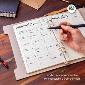 May include: An open planner with the title "Pflanzplan" is being written on with a black pen. The planner is open to a page with a calendar layout, with the months of July, August, and September visible. The text "Jährlich wiederverwendbar Minimalistisch & Übersichtlich" is at the bottom.
