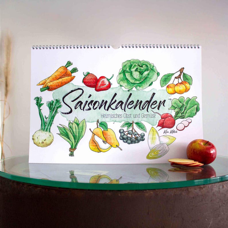 Wandkalender im Querformat mit handgemalten Aquarell-Illustrationen von saisonalem Obst und Gemüse. Der Schriftzug „Saisonkalender“ steht im Mittelpunkt. Daneben liegen frische Tomaten, Rosenkohl, ein Pinsel und ein Farbkasten auf einem Holztisch. Ideal für die Küche oder als Geschenk. Praktisch und dekorativ zugleich. Gedruckt auf hochwertigem Papier. Perfekt für alle, die saisonal und nachhaltig leben möchten.