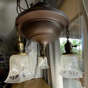 May include: A three-light ceiling fixture featuring a dark bronze-toned metal canopy and base. Three frosted glass shades with etched floral patterns hang from brass-colored sockets, each suspended by a short, twisted metal chain link.