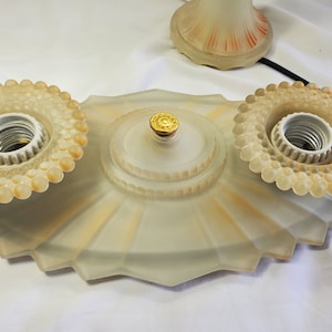 May include: A frosted glass ceiling light fixture with three bulb sockets and a gold-colored center piece. The fixture has a scalloped edge and a beaded design around each socket.
