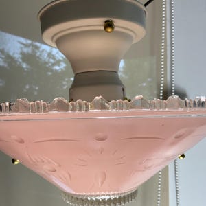 May include: A ceiling light fixture featuring a pale pink frosted glass shade with etched starburst and circular patterns. The shade has a clear, serrated glass rim and is attached to a white metal base with small brass-colored hardware.