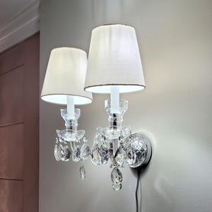 May include: A pair of wall sconces with white fabric shades and crystal accents. The sconces have a silver base and are mounted on a light gray wall. The shades are trimmed with a brown border.