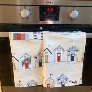 K&ouml;nnte beinhalten: Zwei wei&szlig;e K&uuml;chent&uuml;cher mit einem Strandh&uuml;tten-Design. Die Handt&uuml;cher h&auml;ngen an einer silbernen Backofent&uuml;r. Der Backofen hat ein digitales Display, das "8:16" anzeigt, und der Text "COMPETENCE" ist auf die Backofent&uuml;r gedruckt.