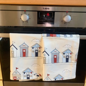 K&ouml;nnte beinhalten: Zwei wei&szlig;e K&uuml;chent&uuml;cher mit einem Strandhaus-Muster. Die Handt&uuml;cher haben blaue und rote Streifen, wei&szlig;e Wolken und kleine rote Herzen. Die Handt&uuml;cher h&auml;ngen an einer Edelstahl-Ofent&uuml;r mit einem digitalen Display, das "88:16" anzeigt. Der Backofen ist mit "AEG Competence" beschriftet.