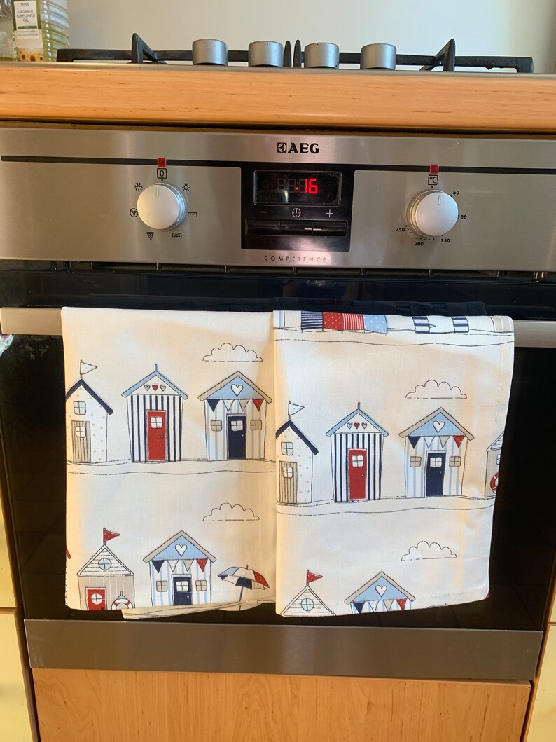 K&ouml;nnte beinhalten: Zwei wei&szlig;e K&uuml;chent&uuml;cher mit einem Strandhaus-Muster. Die Handt&uuml;cher haben blaue und rote Streifen, wei&szlig;e Wolken und kleine rote Herzen. Die Handt&uuml;cher h&auml;ngen an einer Edelstahl-Ofent&uuml;r mit einem digitalen Display, das "88:16" anzeigt. Der Backofen ist mit "AEG Competence" beschriftet.