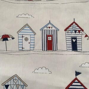 K&ouml;nnte beinhalten: Ein wei&szlig;er Stoff mit einem sich wiederholenden Muster aus blau-wei&szlig; gestreiften Strandh&uuml;tten mit roten D&auml;chern und wei&szlig;en Wolken. Die H&uuml;tten haben rote T&uuml;ren und wei&szlig;e Herzen auf der Vorderseite. In der oberen linken Ecke befindet sich ein wei&szlig;er Schirm mit roter Spitze.