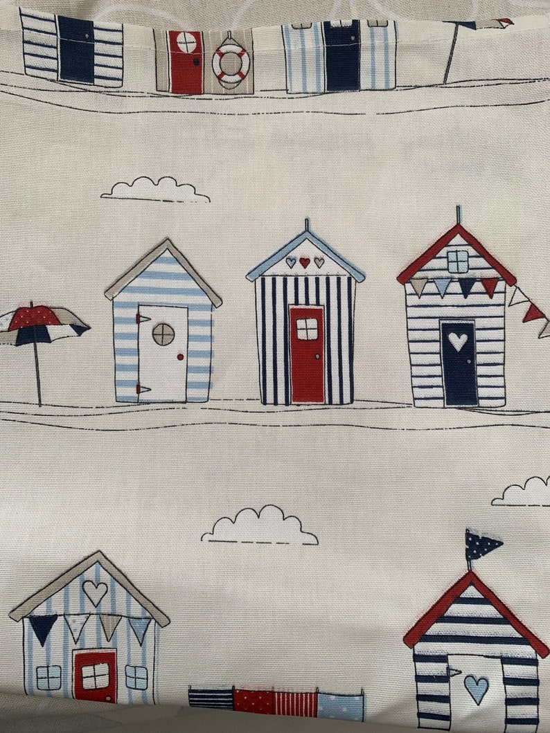 K&ouml;nnte beinhalten: Ein wei&szlig;er Stoff mit einem sich wiederholenden Muster aus blau-wei&szlig; gestreiften Strandh&uuml;tten mit roten D&auml;chern und wei&szlig;en Wolken. Die H&uuml;tten haben rote T&uuml;ren und wei&szlig;e Herzen auf der Vorderseite. In der oberen linken Ecke befindet sich ein wei&szlig;er Schirm mit roter Spitze.