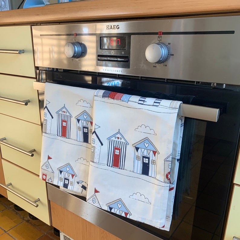 K&ouml;nnte beinhalten: Zwei wei&szlig;e Geschirrt&uuml;cher mit Strandh&uuml;tten-Motiv h&auml;ngen an einer silbernen Backofent&uuml;r. Die Handt&uuml;cher zeigen rote, blaue und wei&szlig;e Strandh&uuml;tten mit Herzen und Flaggen. Der Backofen hat ein digitales Display und ein silbernes Bedienfeld.