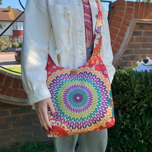 May include: A colorful, multi-colored, mandala-patterned tote bag with a long strap. The bag is worn over the shoulder of a person wearing a white denim jacket and gray pants.