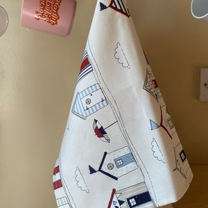 K&ouml;nnte beinhalten: Ein wei&szlig;es Geschirrtuch mit einem blauen und roten Strandh&auml;uschen-Design. Das Geschirrtuch hat oben ein rot-wei&szlig;-blaues Streifenmuster und unten ein blau-wei&szlig;es Streifenmuster. Das Geschirrtuch h&auml;ngt an einem Haken.