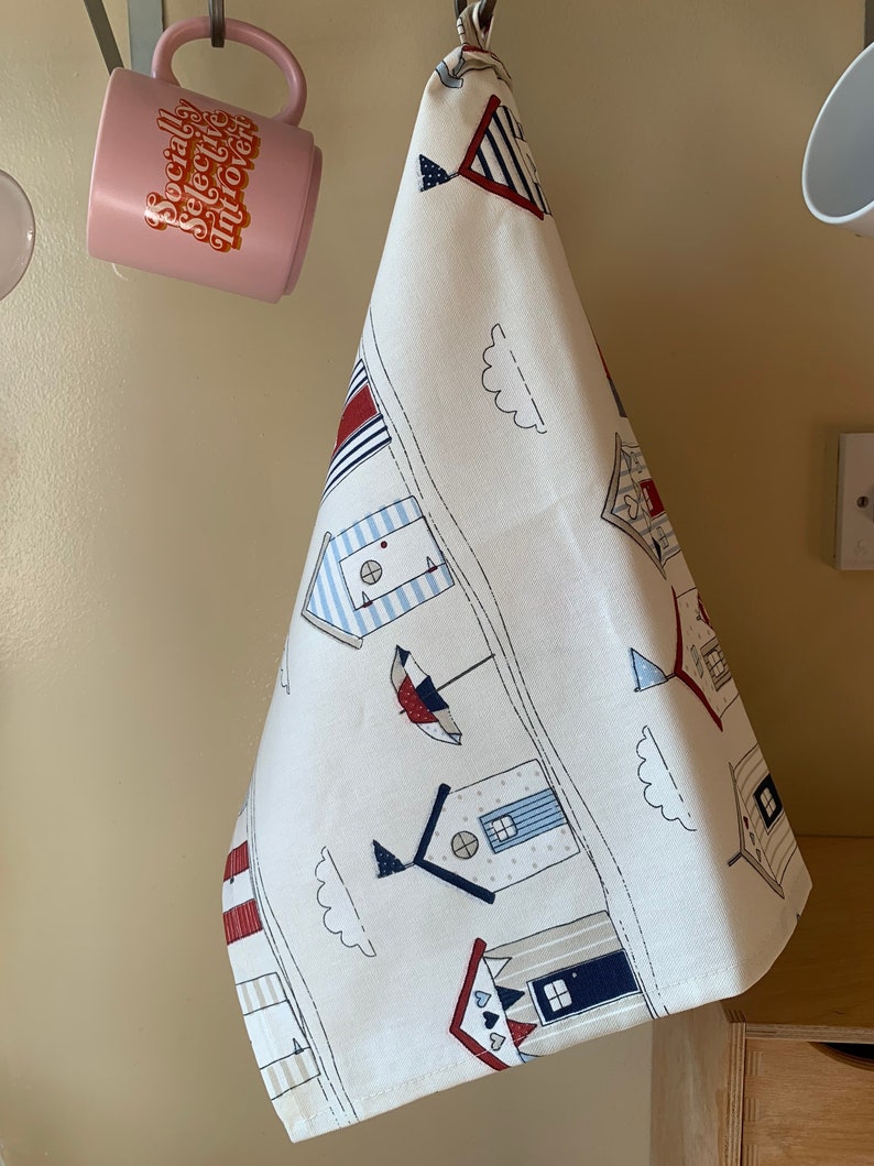 K&ouml;nnte beinhalten: Ein wei&szlig;es Geschirrtuch mit einem blauen und roten Strandh&auml;uschen-Design. Das Geschirrtuch hat oben ein rot-wei&szlig;-blaues Streifenmuster und unten ein blau-wei&szlig;es Streifenmuster. Das Geschirrtuch h&auml;ngt an einem Haken.