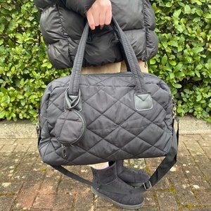 May include: Black quilted duffel bag with a matching round coin purse. The bag has a diamond pattern, top handles, and a detachable shoulder strap. The bag is held in front of a person wearing a black puffer jacket and black boots.