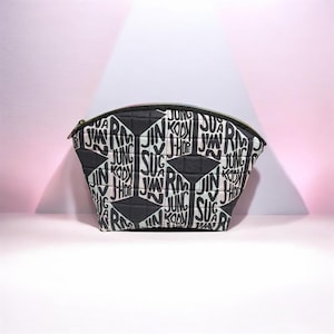 May include: A quilted pouch with a curved top zipper closure, featuring a repeating black and white graphic pattern of names including RM, JIN, SUGA, J-HOPE, JIMIN, V, and JUNG KOOK arranged in a geometric layout with solid black diamond accents.