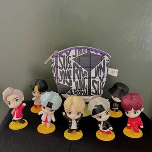 May include: A small, quilted, semi-circular pouch featuring a black and white pattern of repeating names including RM, JIN, and SUGA, with a purple zipper. Seven small, stylized character figurines stand in front, each on a yellow circular base.