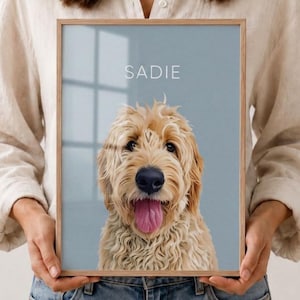 May include: A framed portrait print featuring a digital illustration of a golden-colored doodle dog with curly fur, a black nose, and an open mouth with a pink tongue. The background is a solid light blue, and the name SADIE is printed in white.