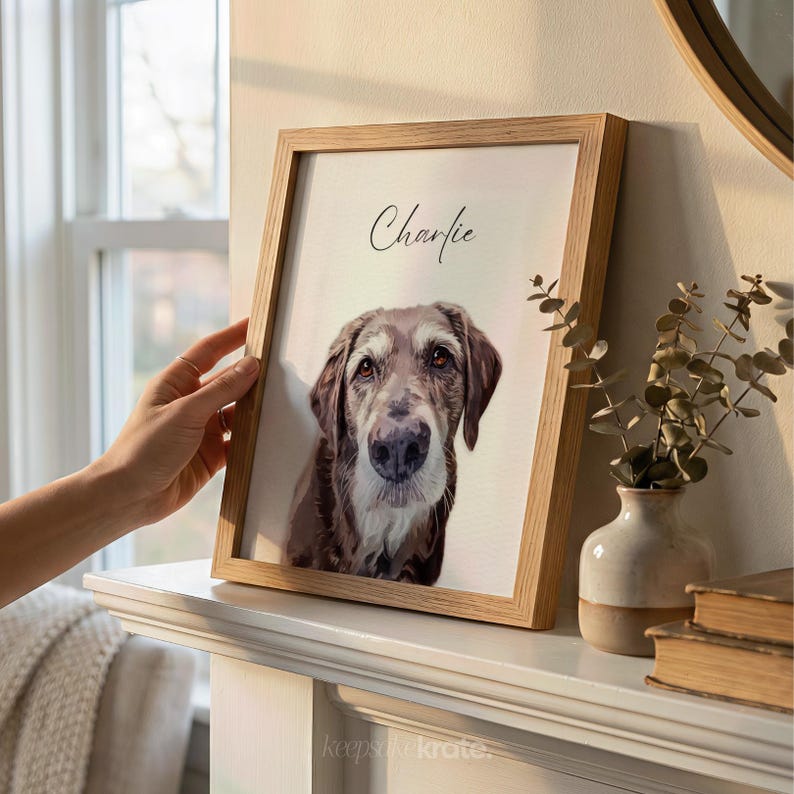 May include: A framed portrait of a dog named Charlie. The artwork features a brown and white dog with a soft, realistic style. The frame is made of light-colored wood. The portrait is displayed on a white mantel.