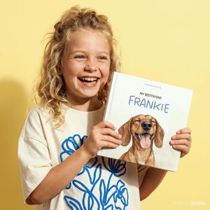 Może przedstawiać: Kwadratowa książka w twardej oprawie zatytułowana My Bestfriend Frankie, przedstawiająca akwarelową ilustrację brązowego jamnika z radosnym wyrazem pyska, otwartym pyskiem i wyciągniętym językiem, wyśrodkowana na białej okładce z niebieskim i czarnym tekstem.