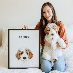 May include: A framed portrait of a dog named Penny, with a black frame and a white background. The dog is white and brown with a fluffy coat. A woman is holding the framed portrait and a live dog that matches the portrait.
