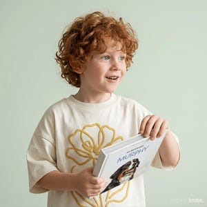 Może przedstawiać: Kremowy t-shirt z kr&oacute;tkim rękawem z dużym, wypukłym złotym konturem kwiatowym z przodu. Koszulkę ma na sobie dziecko z kręconymi rudymi włosami, trzymające białą książkę w twardej oprawie z tytułem My Bestfriend Murphy.