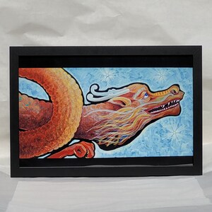 Puede incluir: Una pintura enmarcada de un dragón rojo y naranja con detalles blancos y amarillos. El dragón está volando a través de un cielo azul con copos de nieve blancos.