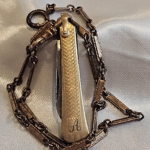 May include: A small, vintage pocket knife with a gold-colored handle and a silver chain. The handle has a textured pattern and the letter "A" engraved on it. The chain is made of rectangular links.