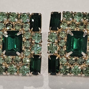 May include: A pair of square-shaped earrings with a gold-colored setting. Each earring is encrusted with emerald green and light green rhinestones. The center stone is an emerald-cut, dark green rhinestone.