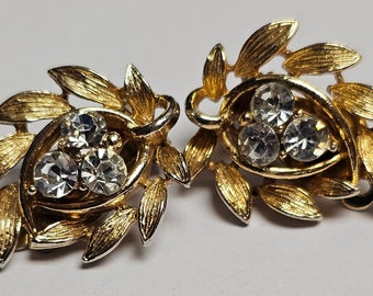 May include: A pair of gold-toned clip-on earrings. Each earring features a cluster of three clear, round rhinestones set within a gold-colored frame, surrounded by textured leaf-shaped accents.