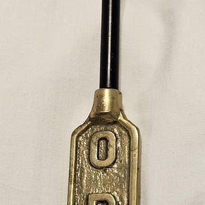 May include: A vertical brass sign with the word OPEN embossed in raised capital letters. The sign is attached to a long, curved black metal handle that features a small brass-colored tip at the end, creating a functional hanging tool.