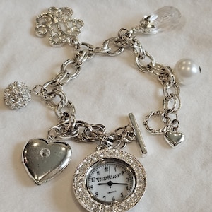 May include: Silver charm bracelet featuring a watch with a crystal-studded bezel, a heart-shaped charm, and other decorative charms. The watch face reads "WHITE LACE". The bracelet has a toggle clasp.