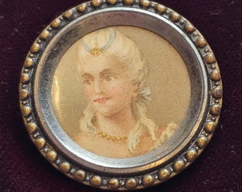 May include: A round antique brooch featuring a portrait of a woman with powdered hair, a pearl necklace, and a delicate dress. The portrait is set within a silver frame and surrounded by a beaded gold border. The background is a deep burgundy.