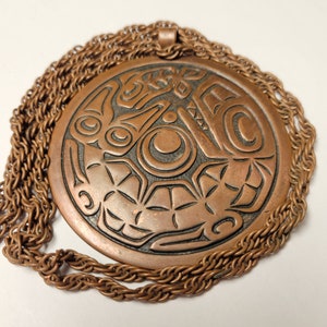 May include: A copper pendant necklace with a circular design featuring intricate carvings of a stylized animal or mythical creature. The pendant is attached to a thick, braided copper chain.