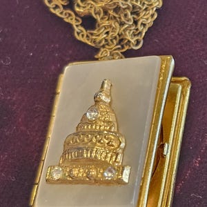 Puede incluir: Collar con colgante de medallón dorado. El colgante rectangular presenta una estructura dorada ornamentada con detalles de cristal transparente sobre un fondo claro. El medallón está abierto, revelando un pequeño espacio interior.