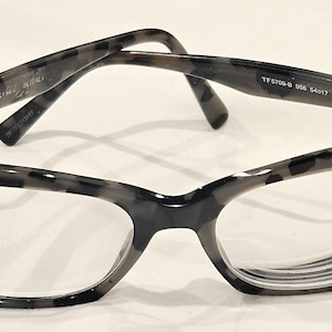 May include: Cat-eye style eyeglasses with a gray and black speckled pattern. The glasses have clear lenses and a silver accent on the frame. The brand name "TOM FORD" is visible on the frame.