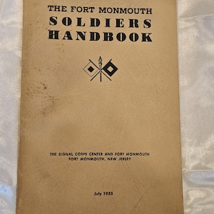 Puede incluir: Un manual antiguo titulado "The Fort Monmouth Soldiers Handbook" con texto negro sobre una cubierta beige. La cubierta incluye el Signal Corps Center y Fort Monmouth, Nueva Jersey, y la fecha de julio de 1953. La cubierta también tiene un gráfico de dos banderas y una antorcha.