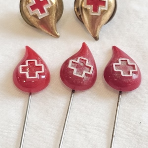 May include: A collection of vintage Red Cross pins and buttons. The image shows two gold-toned buttons with a red cross emblem and three red teardrop-shaped pins with a silver cross design. The pins have long, silver needles.