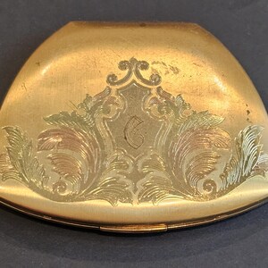 May include: A vintage gold-toned compact with an ornate floral and scroll design. The compact has a curved, triangular shape and a hinged closure. The decorative elements are in shades of gold, green, and copper, adding depth and detail to the surface.