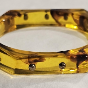 May include: A hexagonal amber-colored bangle bracelet with dark brown marbling. The bracelet has silver-colored studs along the edges. The jewelry is a vintage accessory.