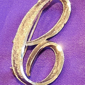 May include: A gold-toned metal brooch in the shape of the letter "B" against a purple background. The brooch has a textured surface and a pin closure on the back. The letter is in a cursive script.
