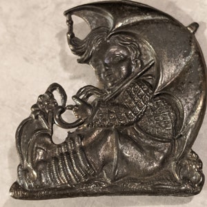 May include: A silver-toned brooch featuring a detailed relief of a person under an umbrella. The person is wearing a patterned garment and holding a violin. The brooch has an antique, vintage aesthetic.