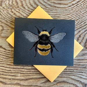 Pode incluir: Um cartão de felicitações com uma ilustração detalhada de um zangão com listras pretas e amarelas e asas translúcidas. O cartão é cinza escuro e repousa sobre um envelope amarelo, sobre um fundo de madeira.