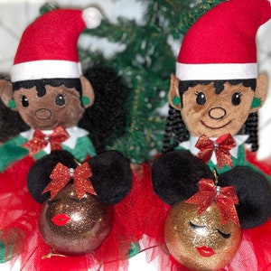 May include: Two elf dolls with black hair and brown skin wearing red and green elf outfits and holding gold and brown glitter ornaments with painted faces. The ornaments are decorated with red bows and glitter.