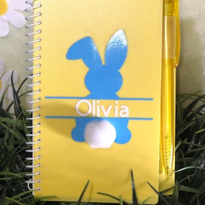 Puede incluir: Cuaderno amarillo de espiral con una silueta de conejo azul y el nombre "Olivia" impreso en él. El cuaderno viene con un bolígrafo amarillo.